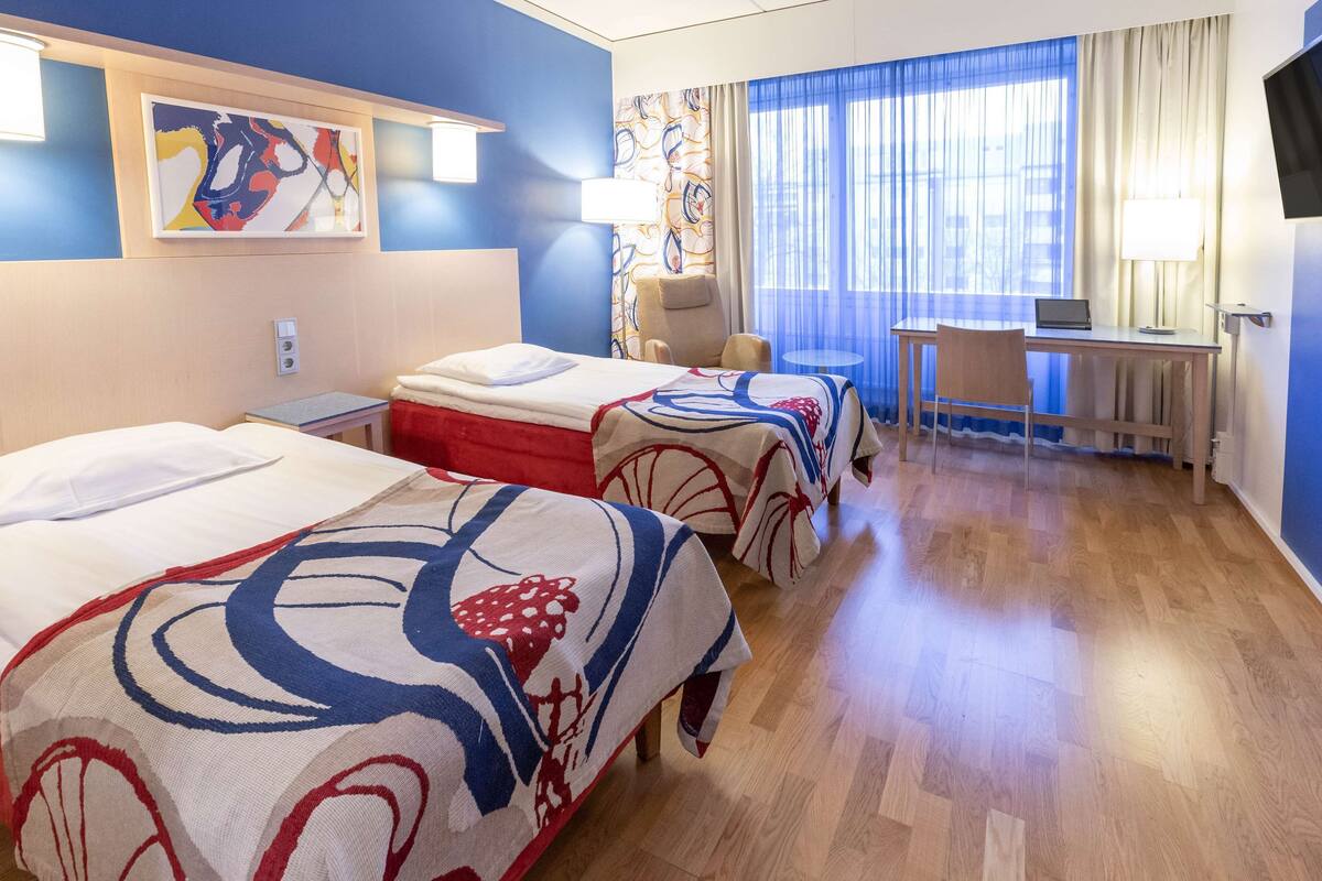 Habitació d'hotel amb dos llits, una taula de treball, una cadira, una televisió i una finestra amb cortines.