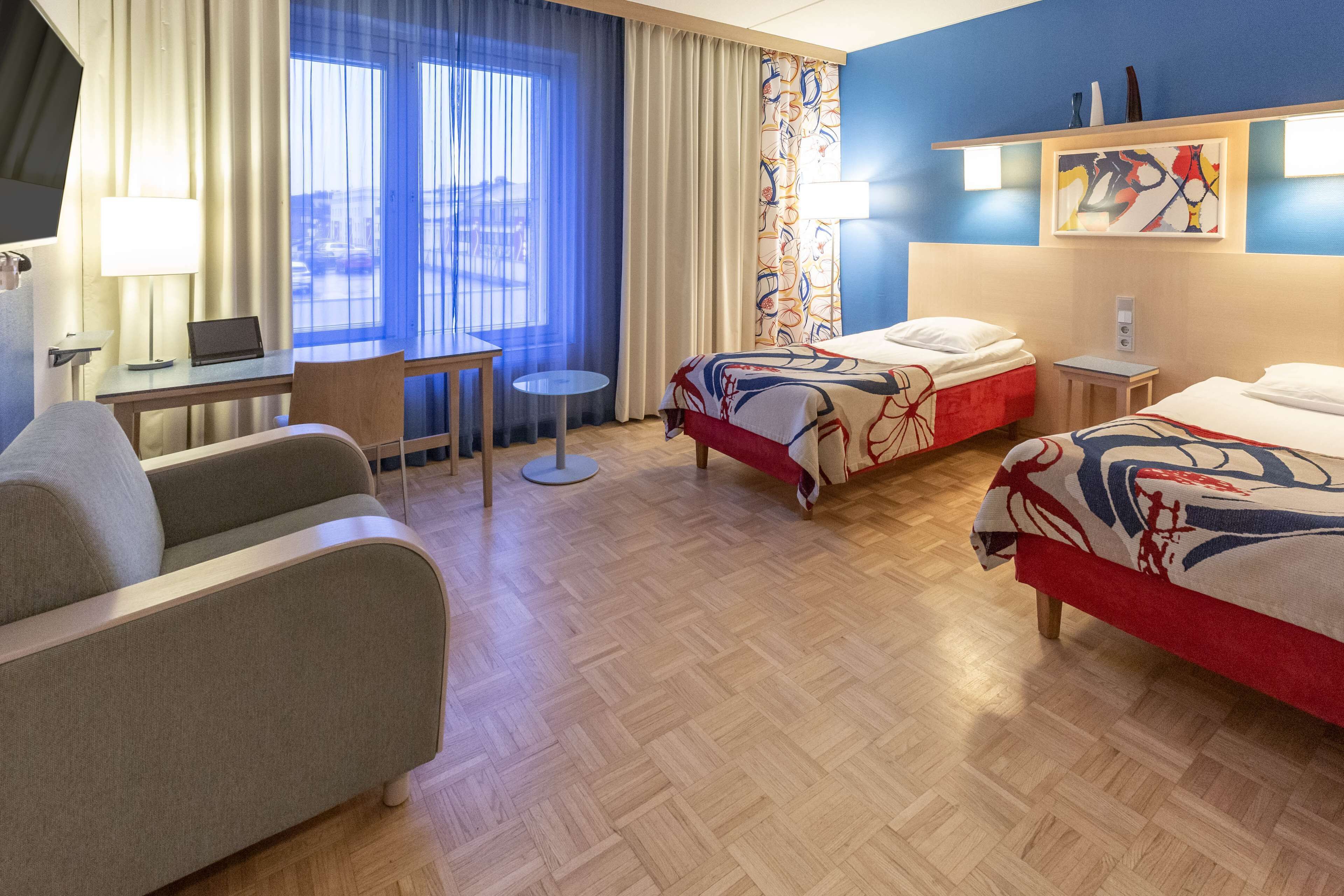 Habitación de hotel con dos camas, un escritorio, una silla, una mesita y una ventana con cortinas.