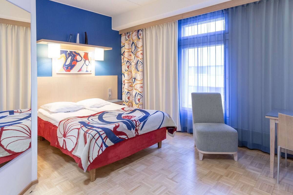 Habitació d'hotel amb llit, cadira, escriptori i finestra amb cortines.