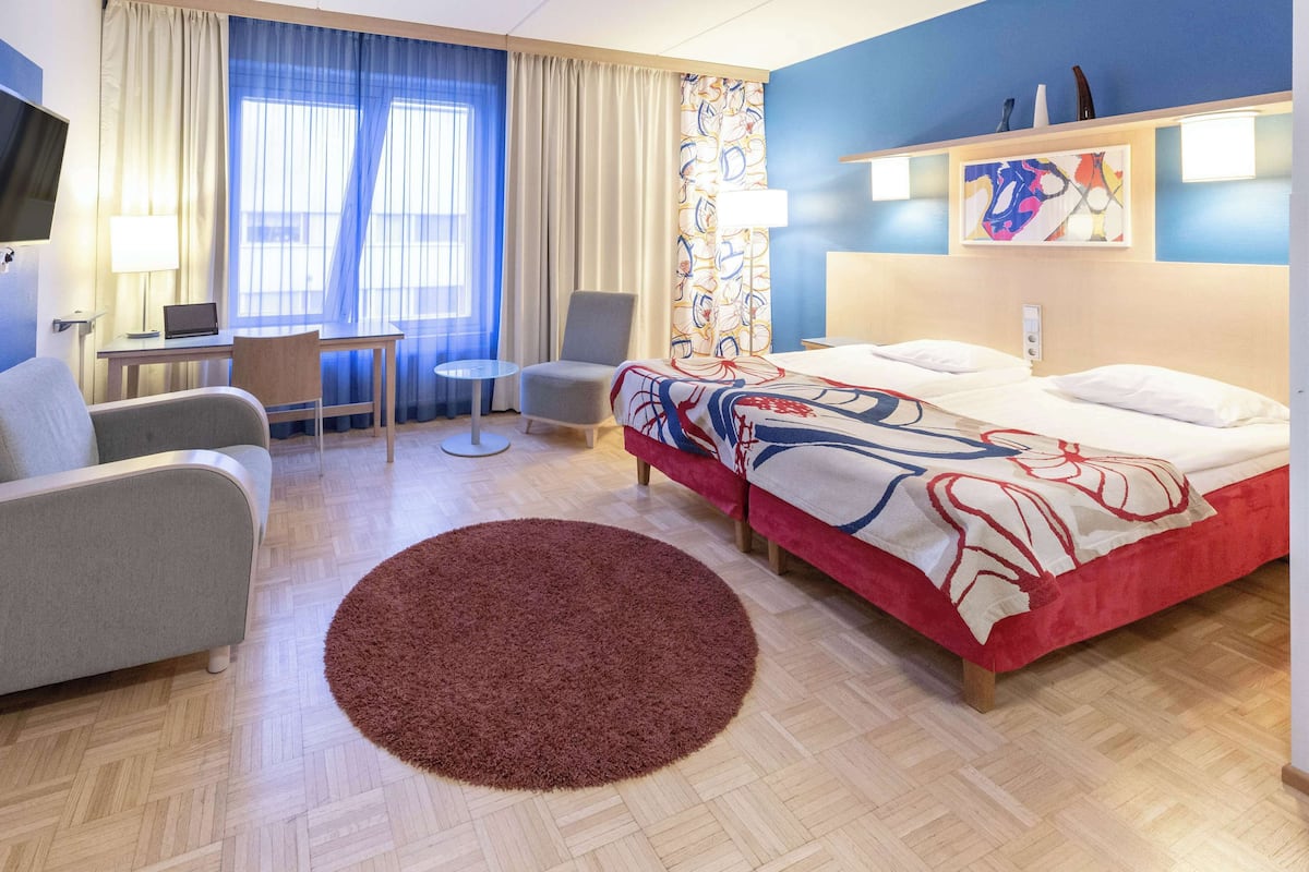 Una habitació d'hotel amb un llit, dues cadires, un escriptori amb un ordinador portàtil, una catifa rodona i una finestra gran amb cortines.