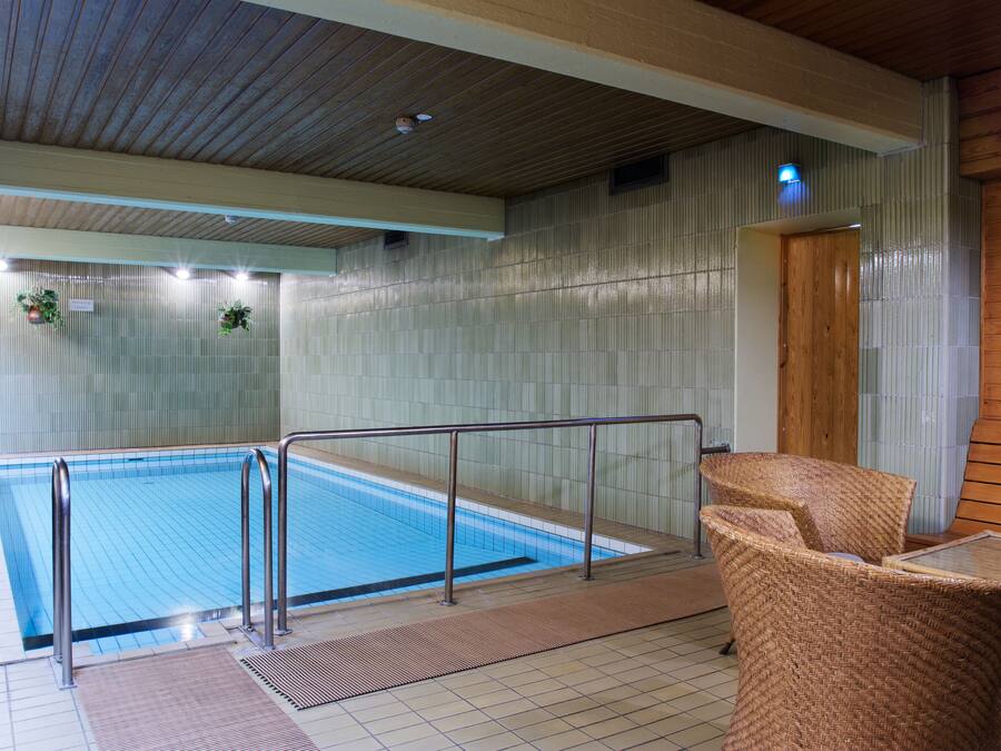 An indoor swimming pool with a wooden ceiling, metal railings, and wicker chairs.