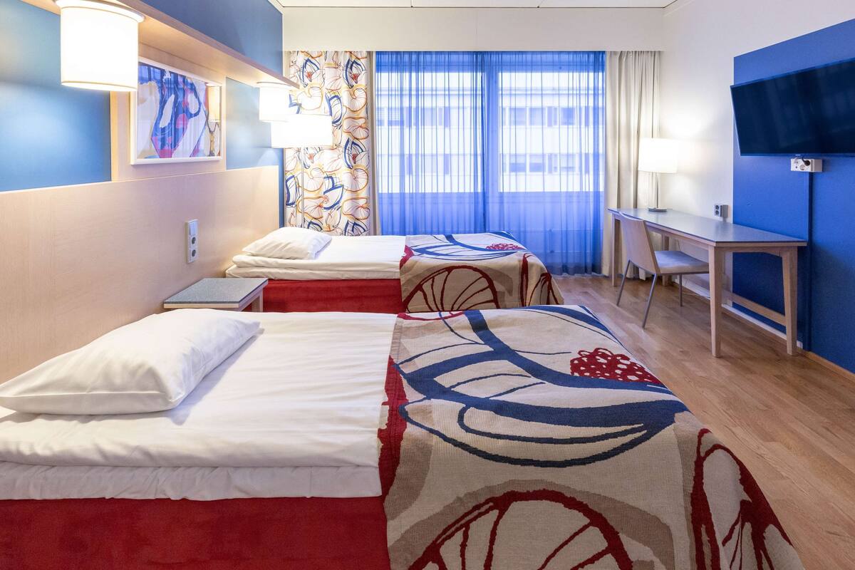 Habitació d'hotel amb dos llits, una taula de treball, televisió i finestra amb cortines.