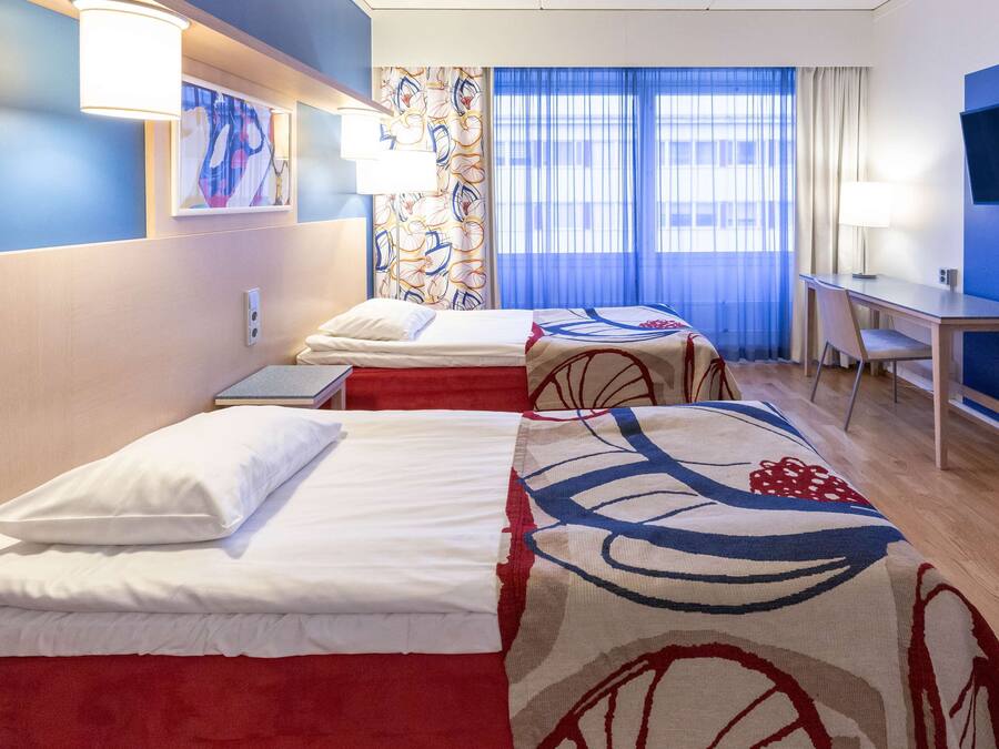 Quarto de hotel com duas camas, uma mesa, uma televisão e uma janela com cortinas.