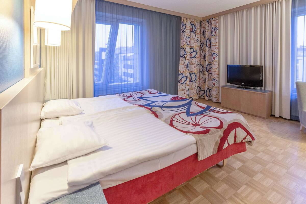 Habitació d'hotel amb un llit, una televisió sobre un suport, una finestra amb cortines i vista a edificis a l'exterior.