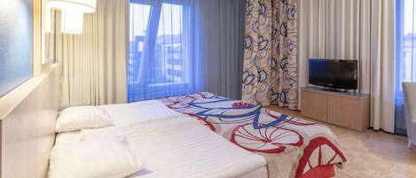 Habitación de hotel con cama, televisor en un mueble bajo, ventana con cortinas y vistas a edificios.