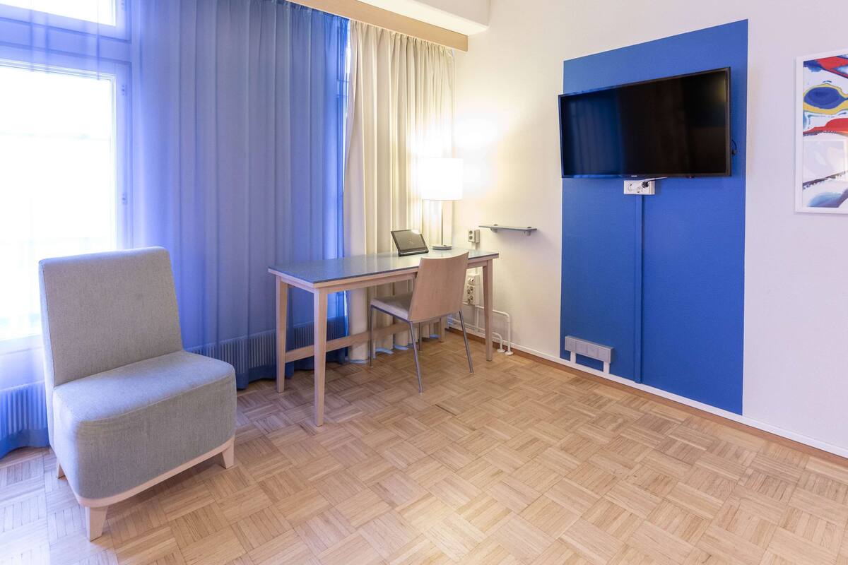 Habitació d'hotel amb sòl de fusta, escriptori amb un ordinador portàtil, una cadira, un televisor fixat a una paret blava i una finestra amb cortines.