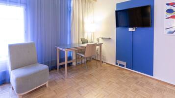 A hotel room with a wooden floor, a desk with a laptop, a chair, a TV mounted on a blue wall, and a window with curtains.