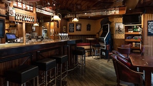 Una zona de bar con suelo de madera, taburetes, una barra con botellas y un televisor montado en la pared.