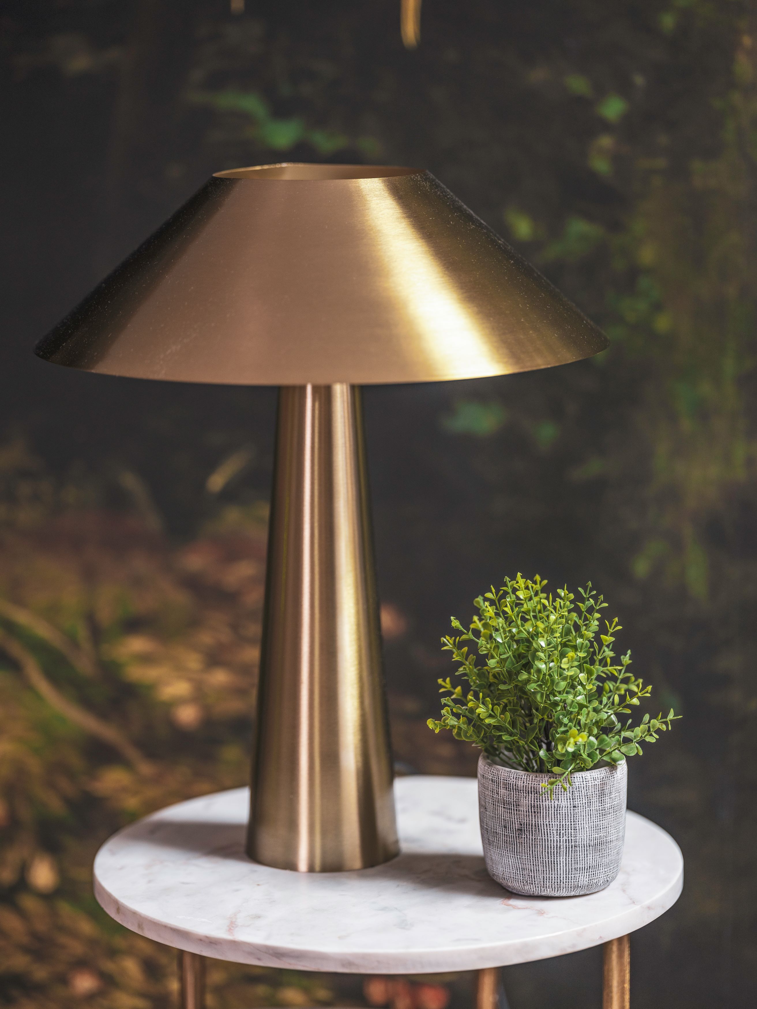 A metallic table lamp with a conical shade, a small potted plant, and a marble table against a blurred background.