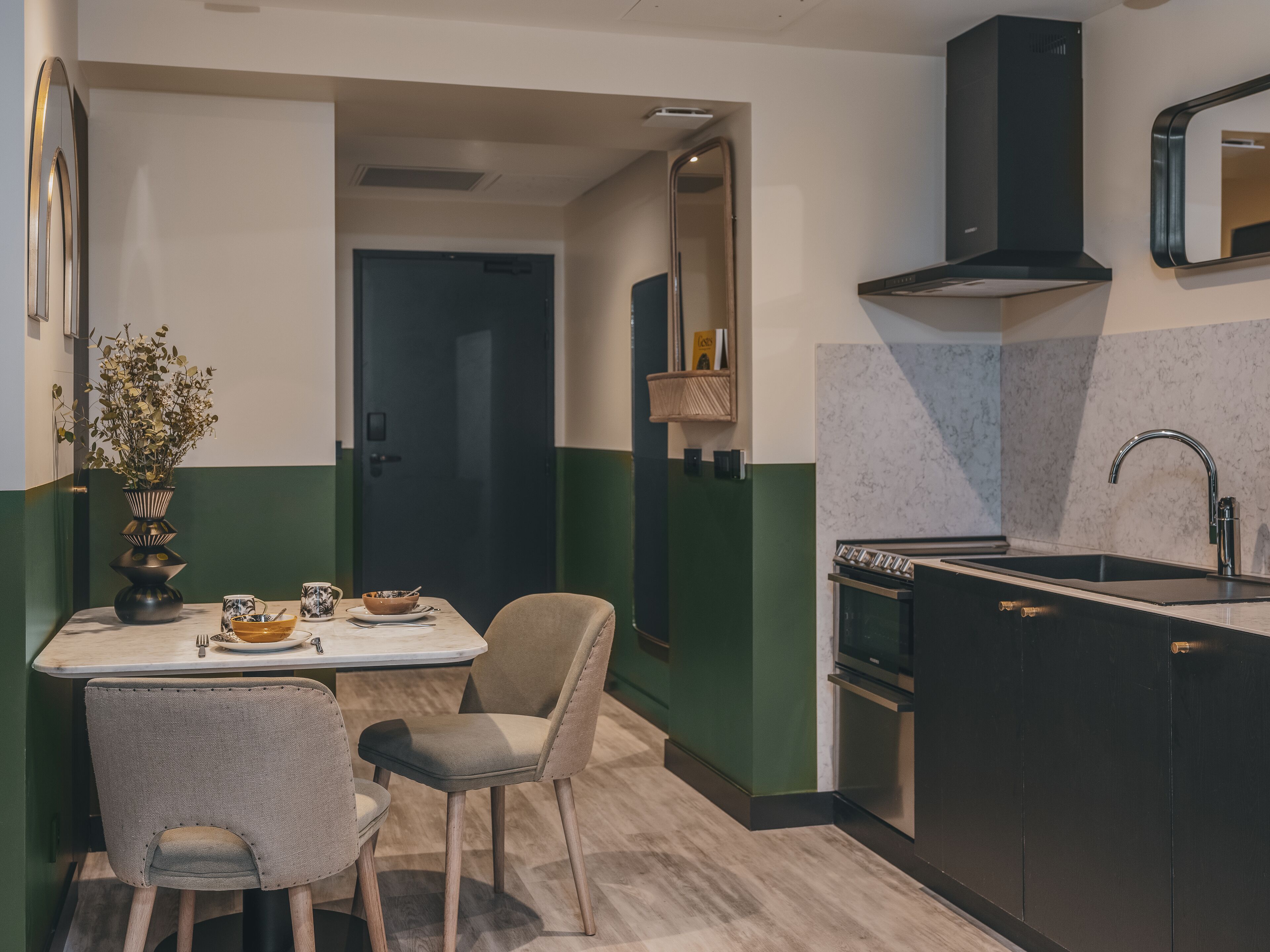 A modern kitchen with a dark countertop, a built-in oven, and a black range hood. There is a dining area with a table and two chairs.