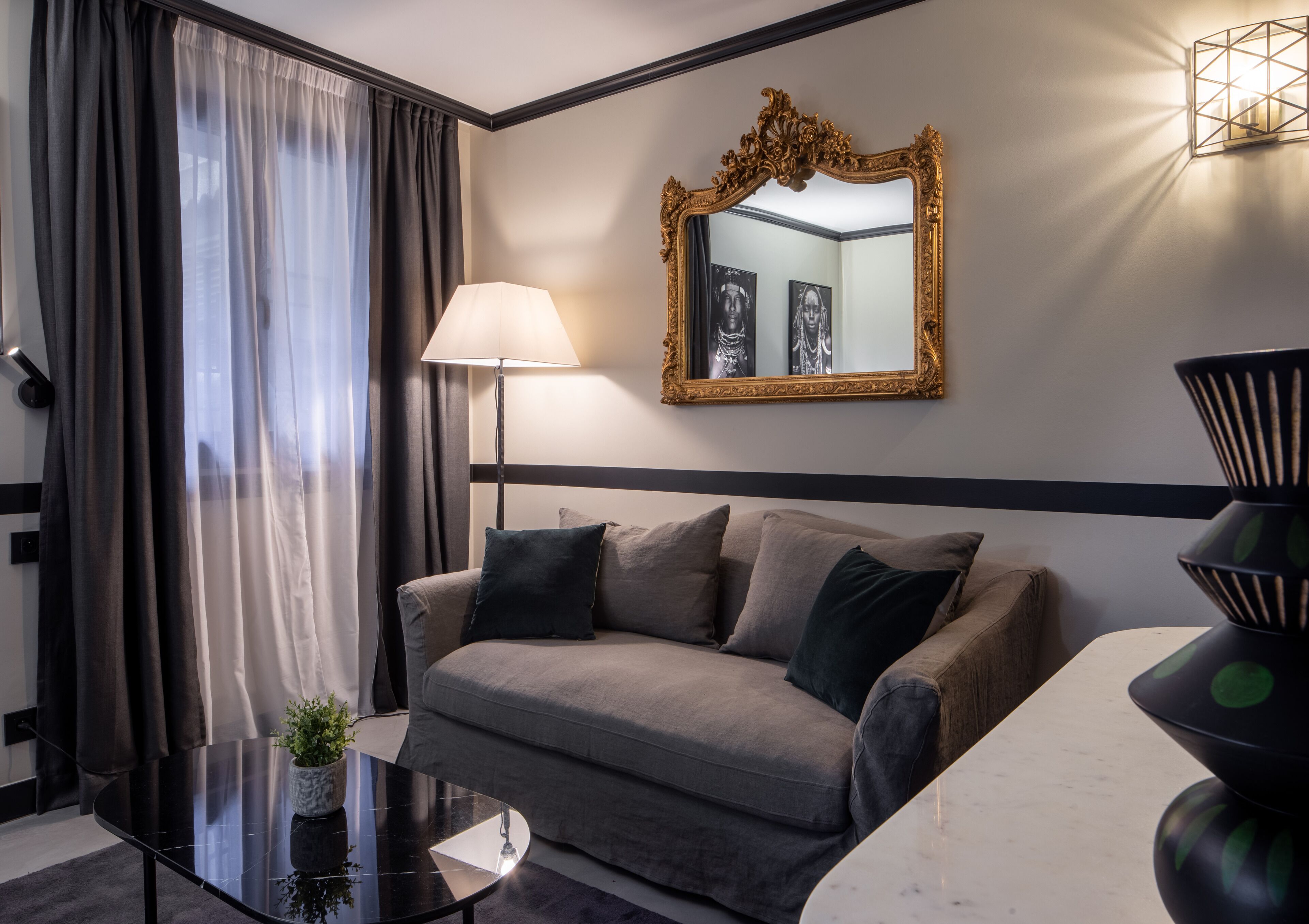 Habitació d'hotel moderna amb un sofà gris, una taula de cafè negra, una làmpada de peu, un mirall gran i un vas decoratiu.