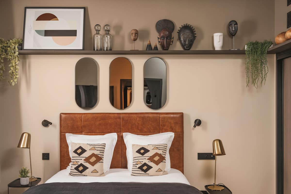 A bedroom with a brown headboard, two bedside tables with lamps, a bed with patterned pillows, three wall mirrors, and decorative items on a shelf.