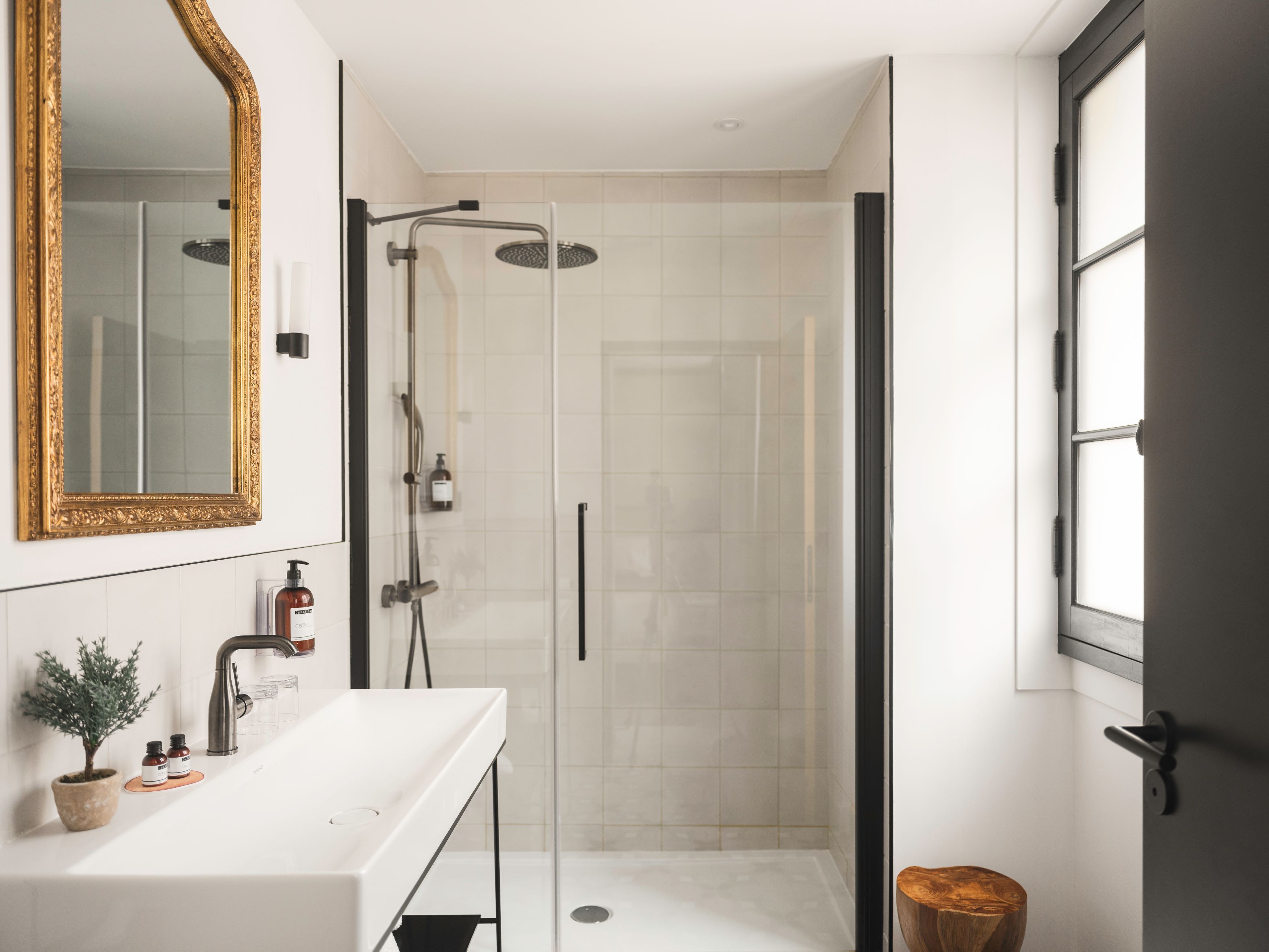 A modern bathroom with a glass-enclosed shower, a white sink with a mirror, and a wooden stool.