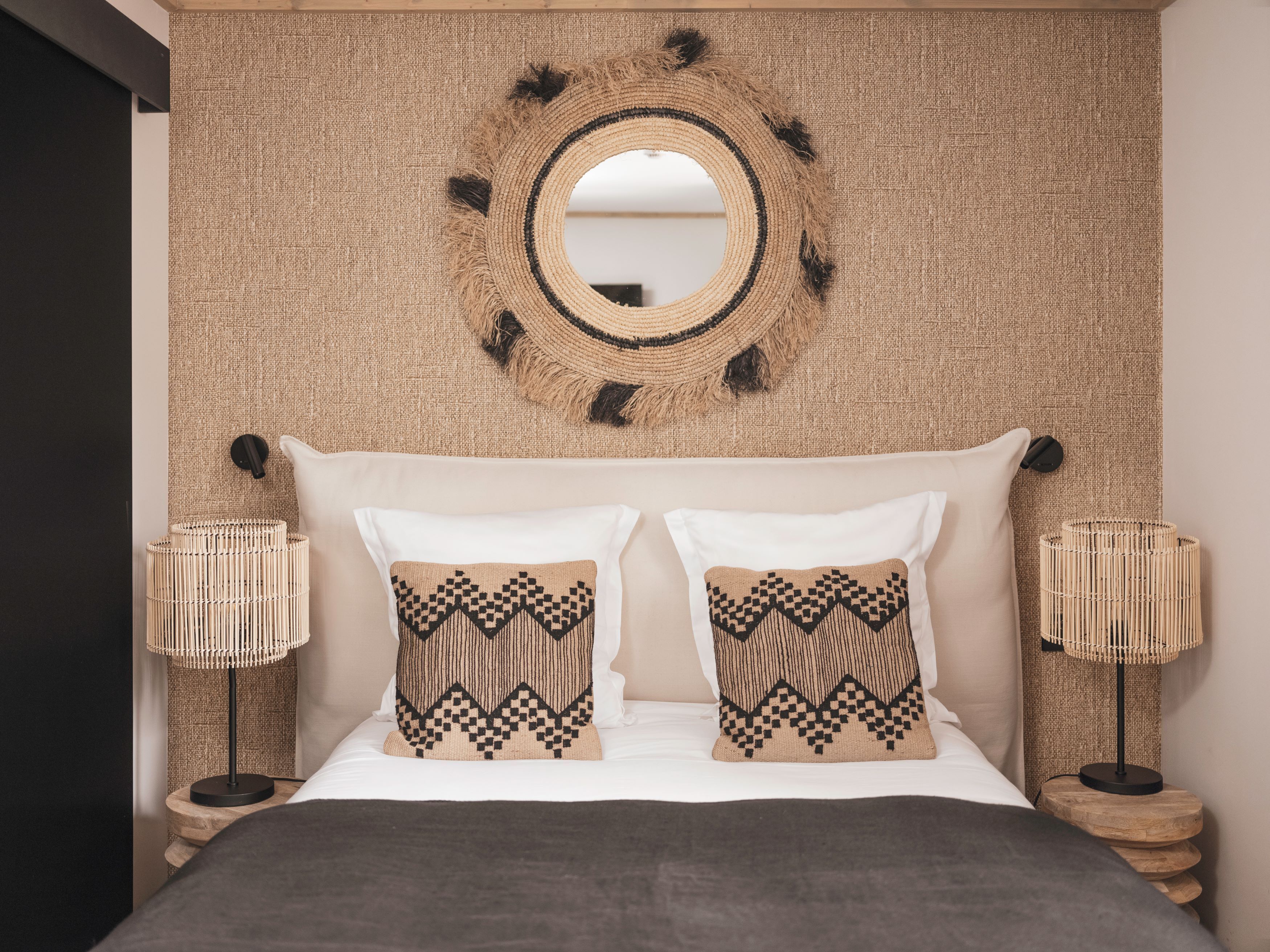 A bed with two pillows, a round mirror, and two lamps on wooden nightstands.