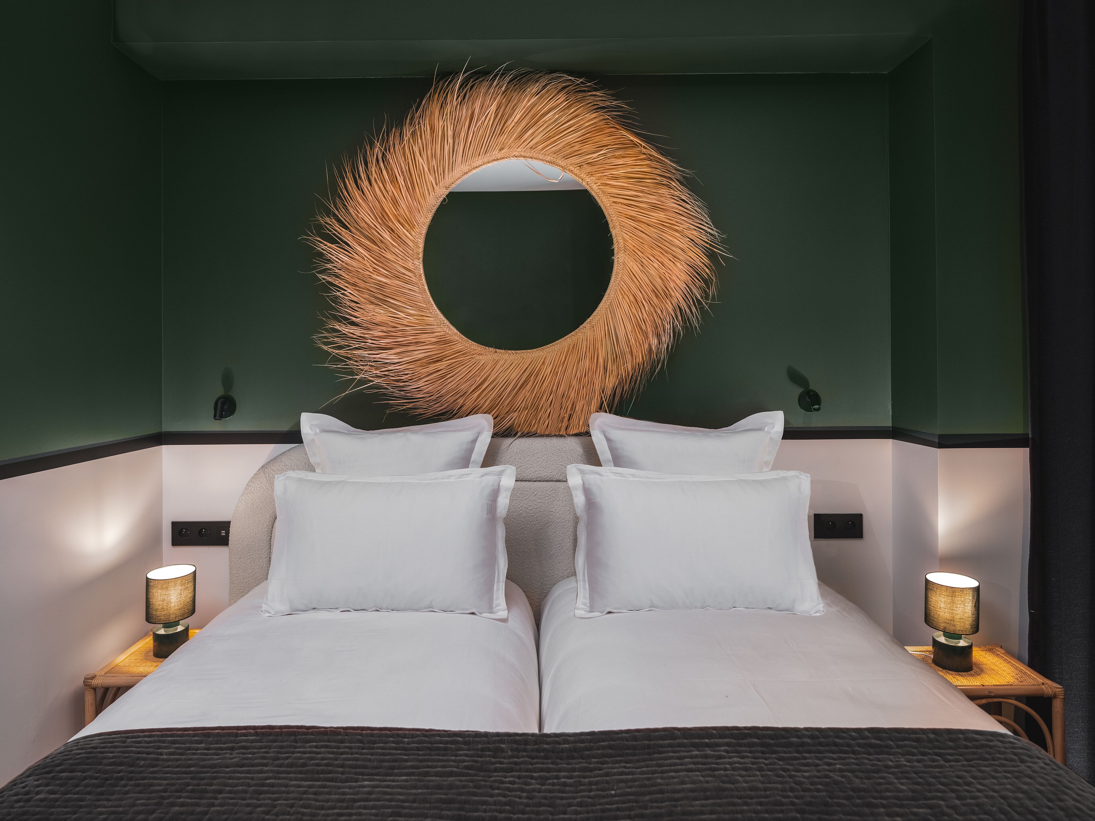 Una cama con ropa blanca, dos lámparas de noche y un espejo grande de forma circular con un marco texturizado.