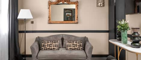Une chambre d’hôtel moderne avec un canapé gris, une table basse ronde noire, un grand miroir décoratif et une petite table d’appoint agrémentée de vases décoratifs.