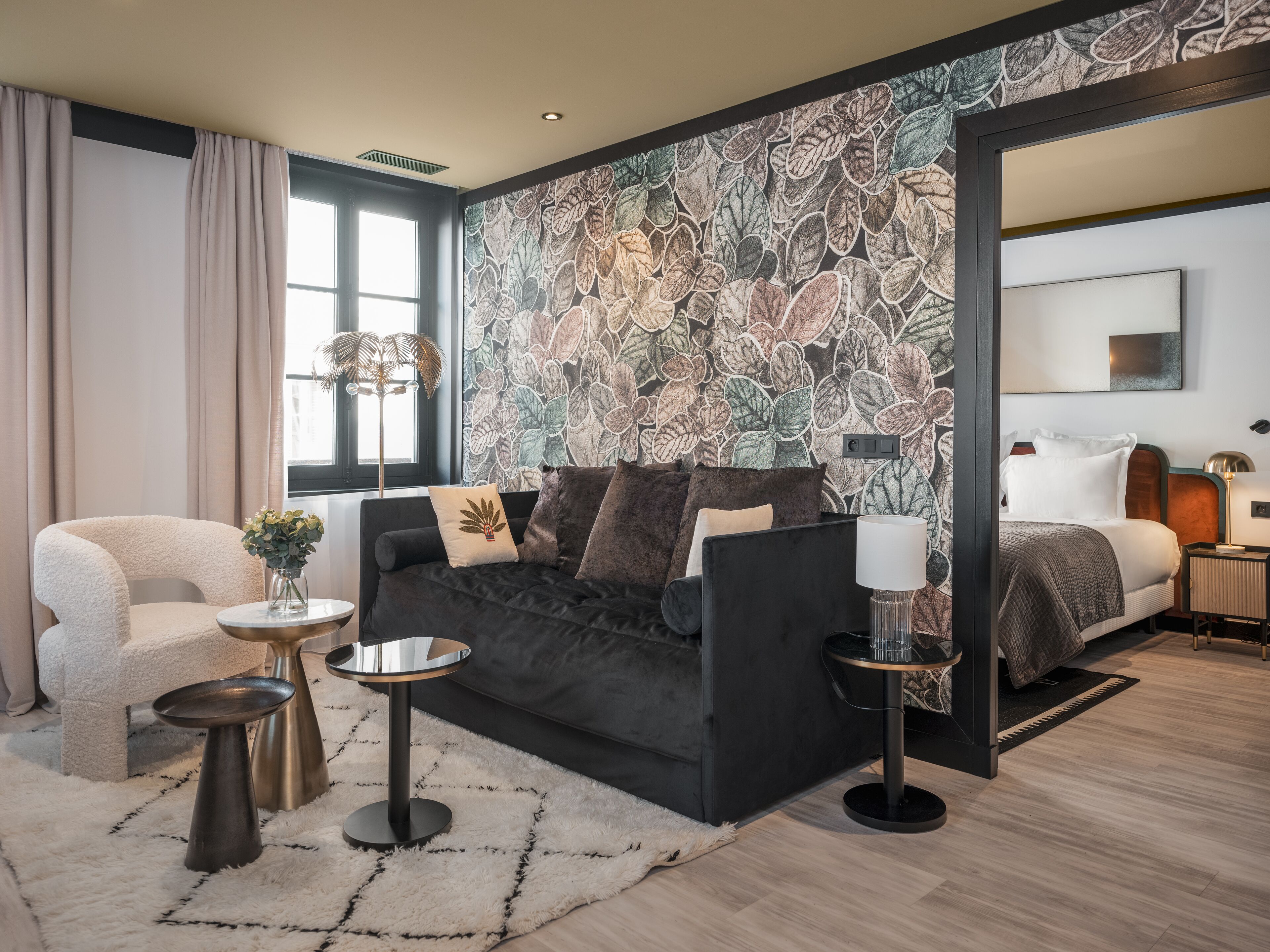 Una habitación de hotel moderna con un sofá negro, una mesita redonda auxiliar, una pequeña mesa redonda, una lámpara de pie y una cama con ropa de cama blanca.