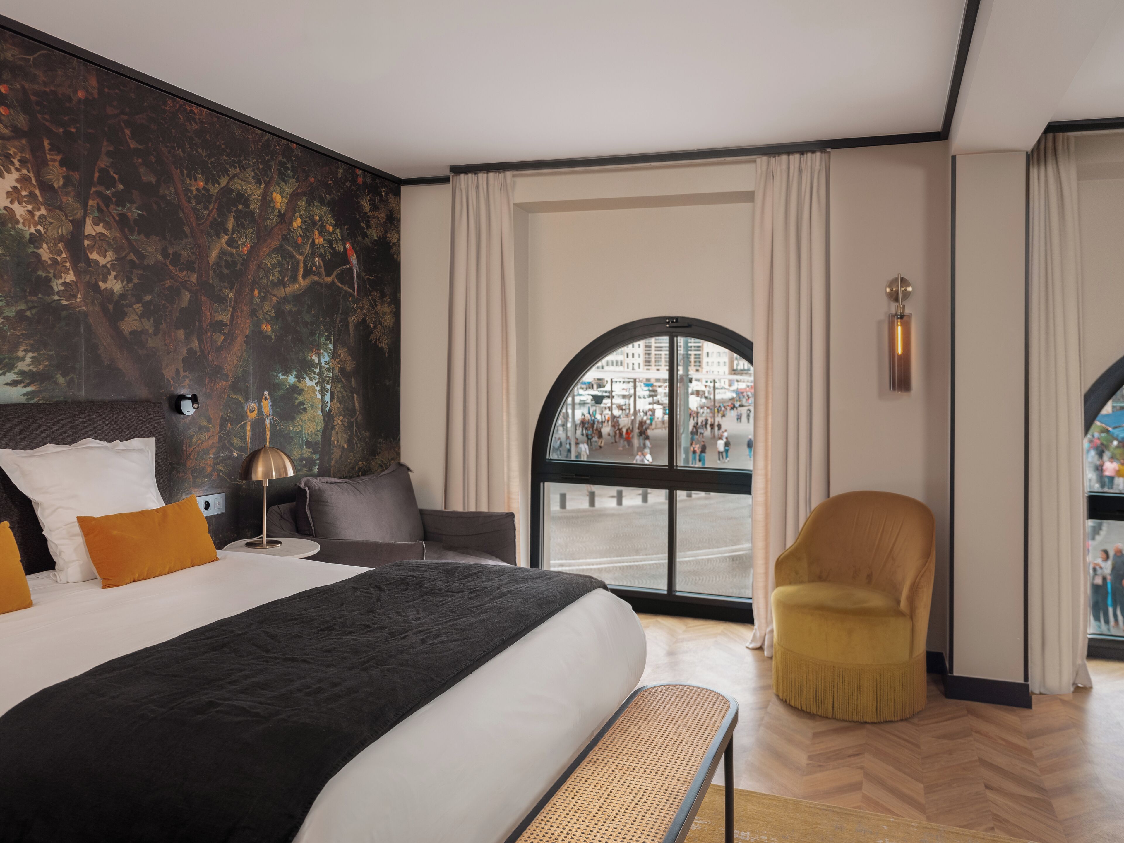 Una habitación de hotel moderna con una cama grande, un banco y vistas a una calle concurrida, además de un mural en la pared.