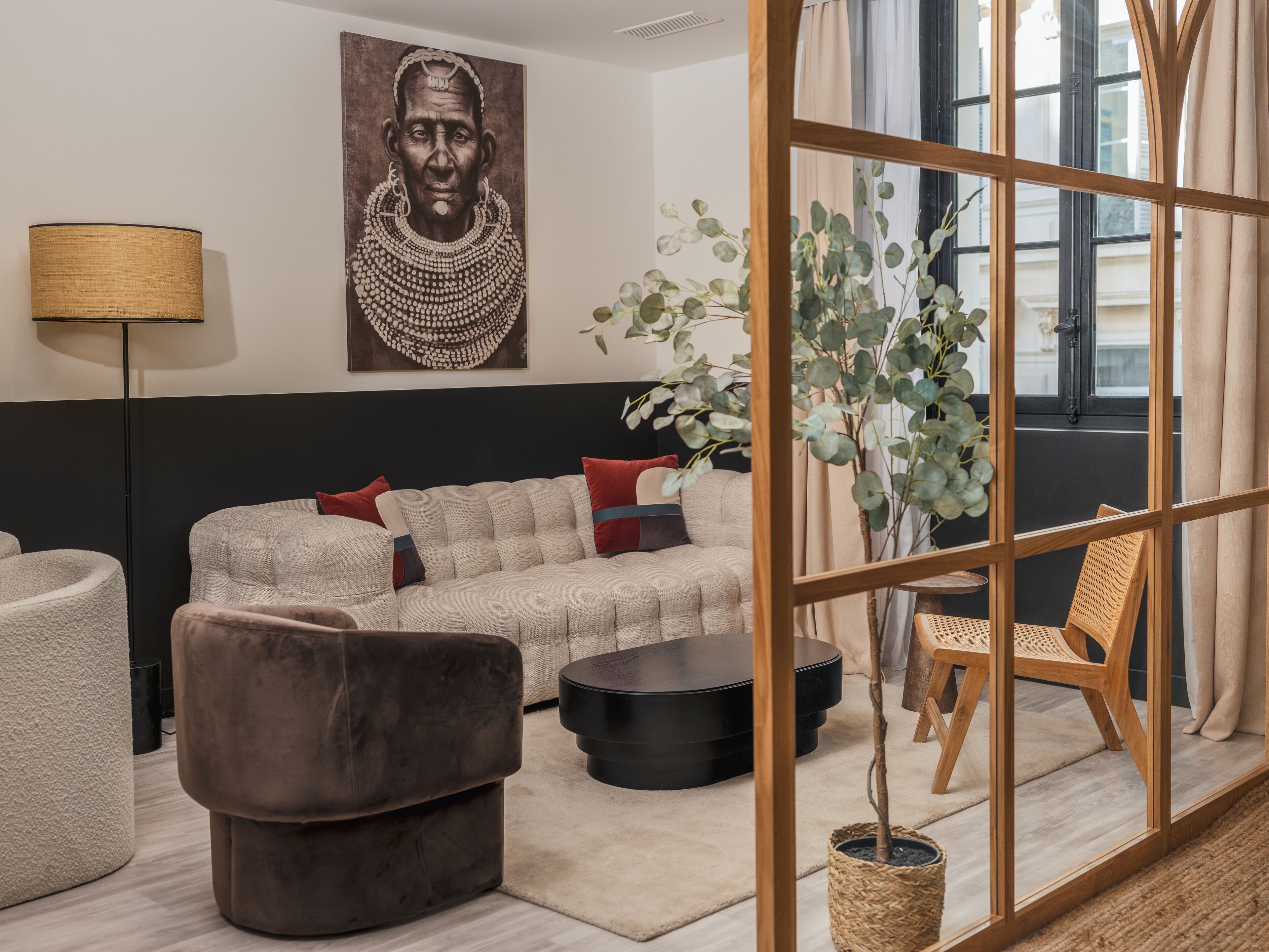 A modern living room with a sectional sofa, a brown armchair, a round black coffee table, and a wooden chair. There is a large painting on the wall, a floor lamp, and a potted plant.