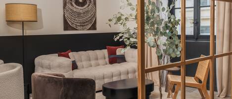 A modern living room with a sectional sofa, a brown armchair, a round black coffee table, and a wooden chair. There is a large painting on the wall, a floor lamp, and a potted plant.