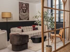 Un salon moderne avec un canapé d’angle, un fauteuil marron, une table basse ronde noire et une chaise en bois. Un grand tableau orne le mur, ainsi qu’un lampadaire et une plante en pot.