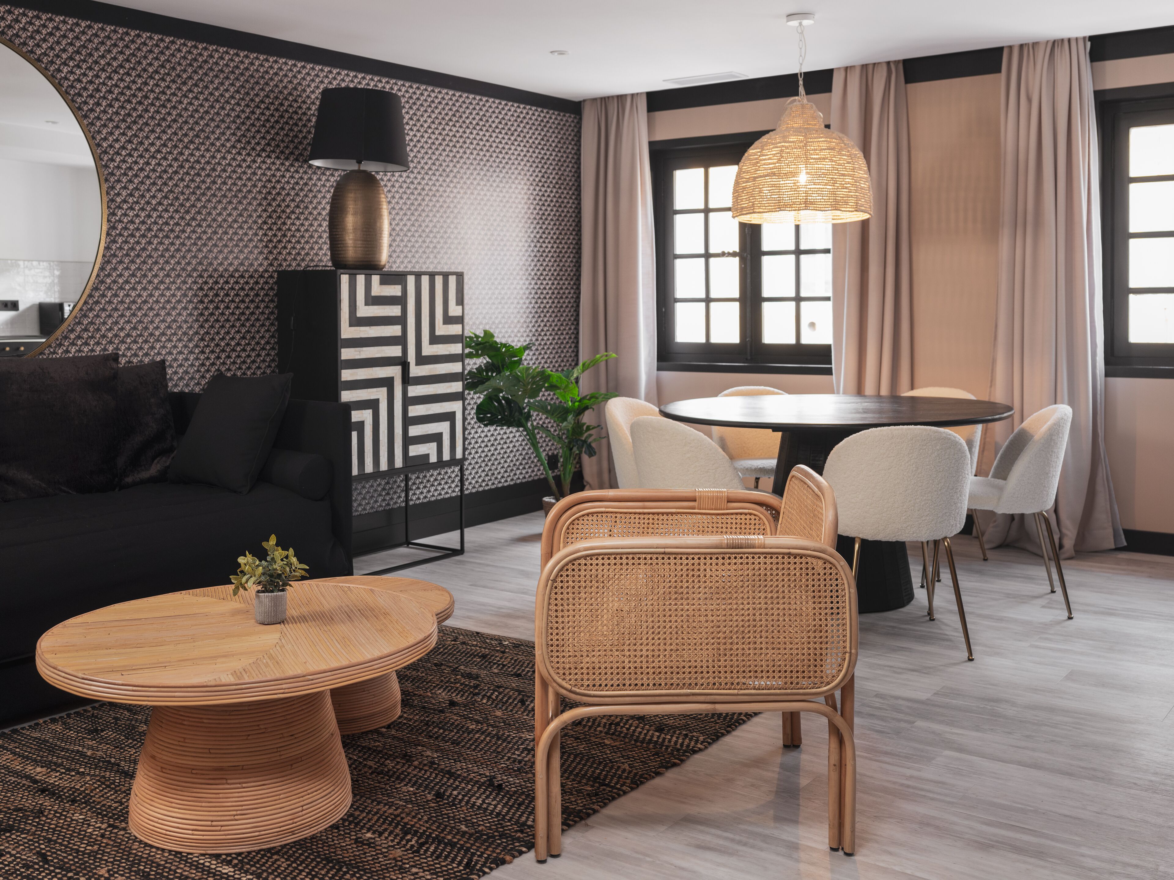 Un salon moderne avec une table de salle à manger ronde, des meubles en osier, un grand miroir et un mur décoré d’un motif.