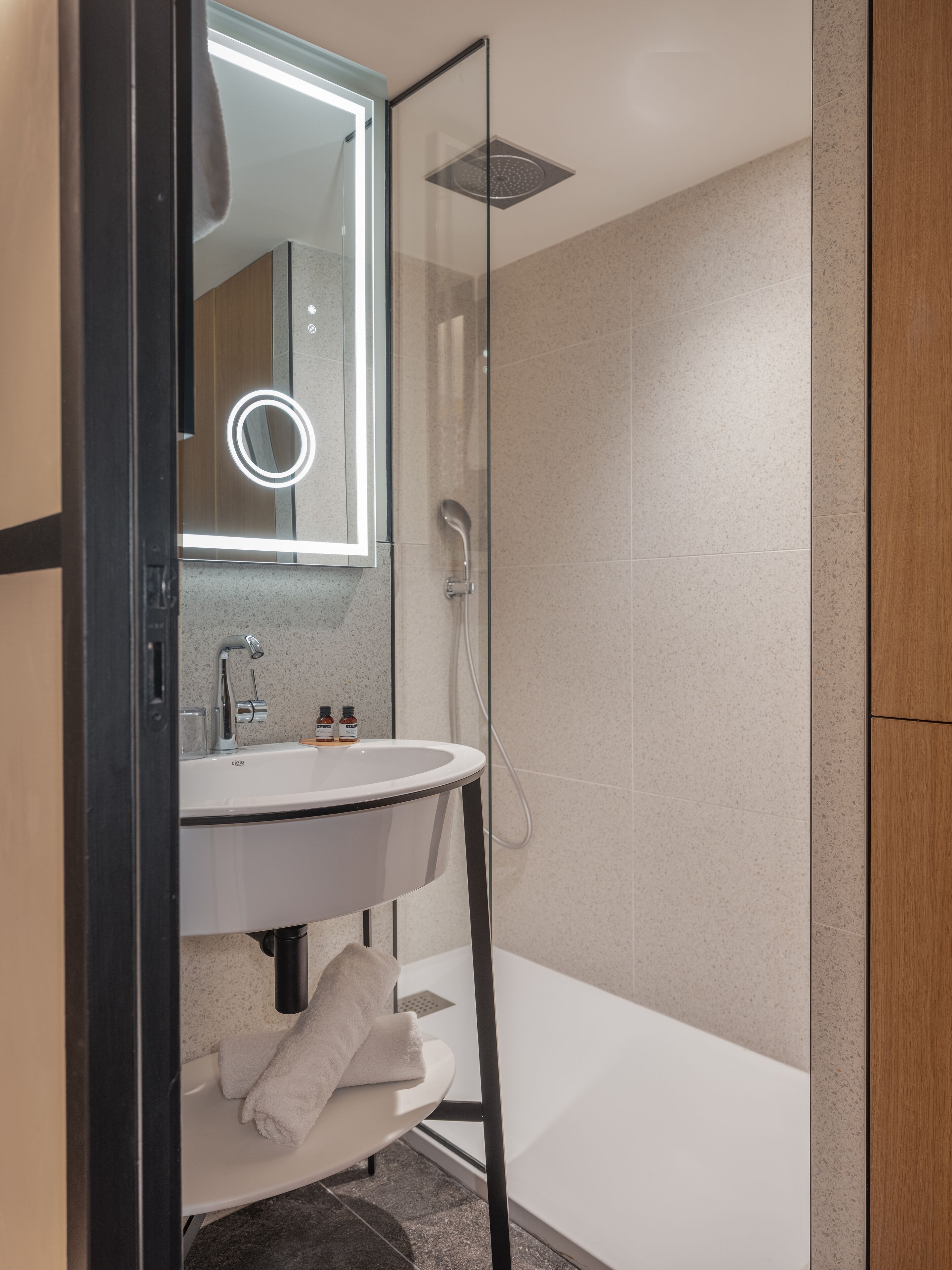 Un baño moderno con ducha de vidrio, lavamanos blanco y espejo con luz circular.