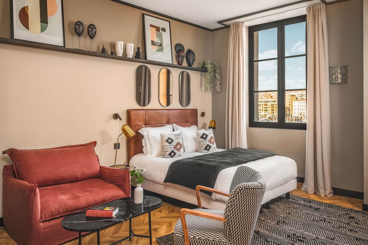 A modern hotel room with a bed, a red sofa, a patterned armchair, a small table, and a large window with a city view.