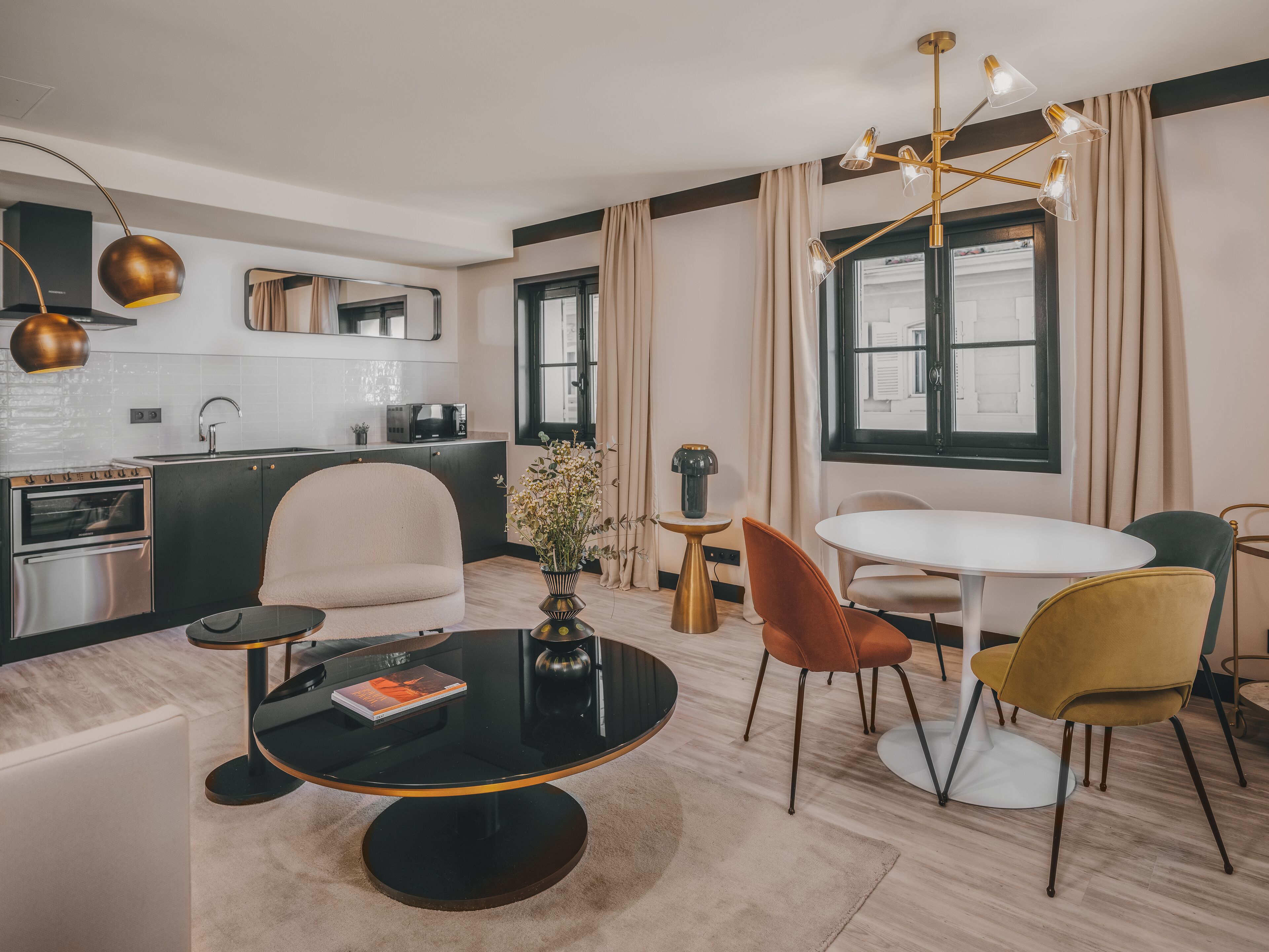 A modern living room with a round black coffee table, beige armchairs, a white dining table, and a kitchen in the background.