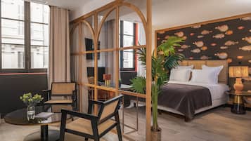 Quarto de hotel com uma grande janela, cama com roupa branca, cabeceira de madeira, mesa de cabeceira com abajur, planta em vaso e vista para a paisagem urbana.