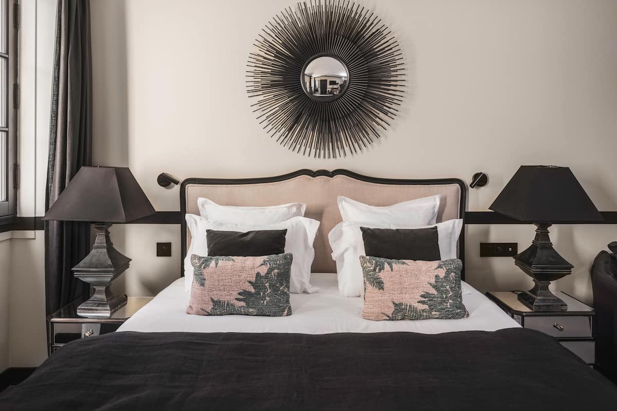 A neatly made bed with a black and white headboard, two bedside lamps, and a wall-mounted mirror with a sunburst design.