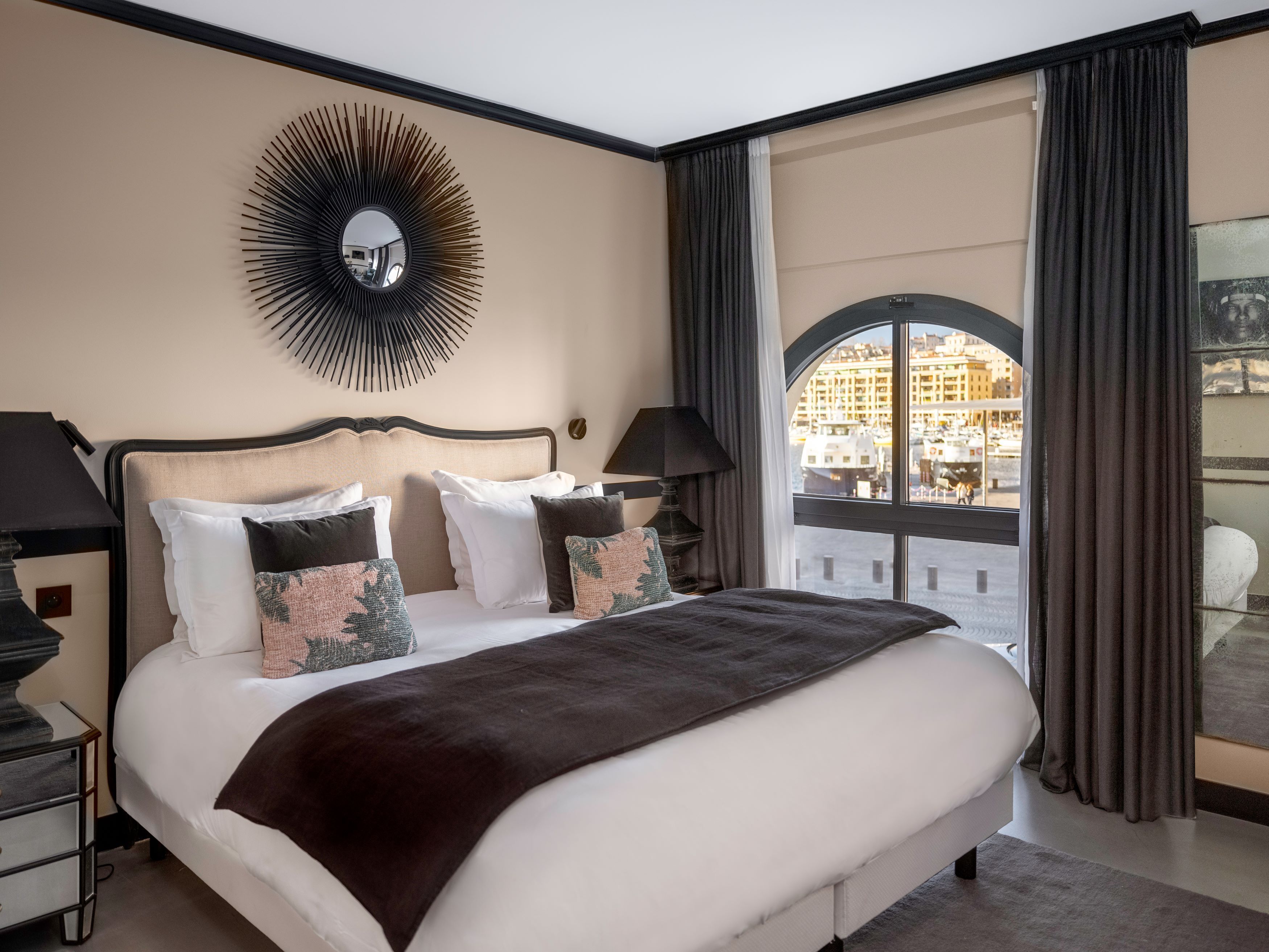 Hotelkamer met een groot bed, een ronde spiegel, een nachtkastje, een lamp en een uitzicht op een stadsbeeld door een groot raam.