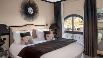 A hotel room with a large bed, a round mirror, a nightstand, a lamp, and a view of a cityscape through a large window.