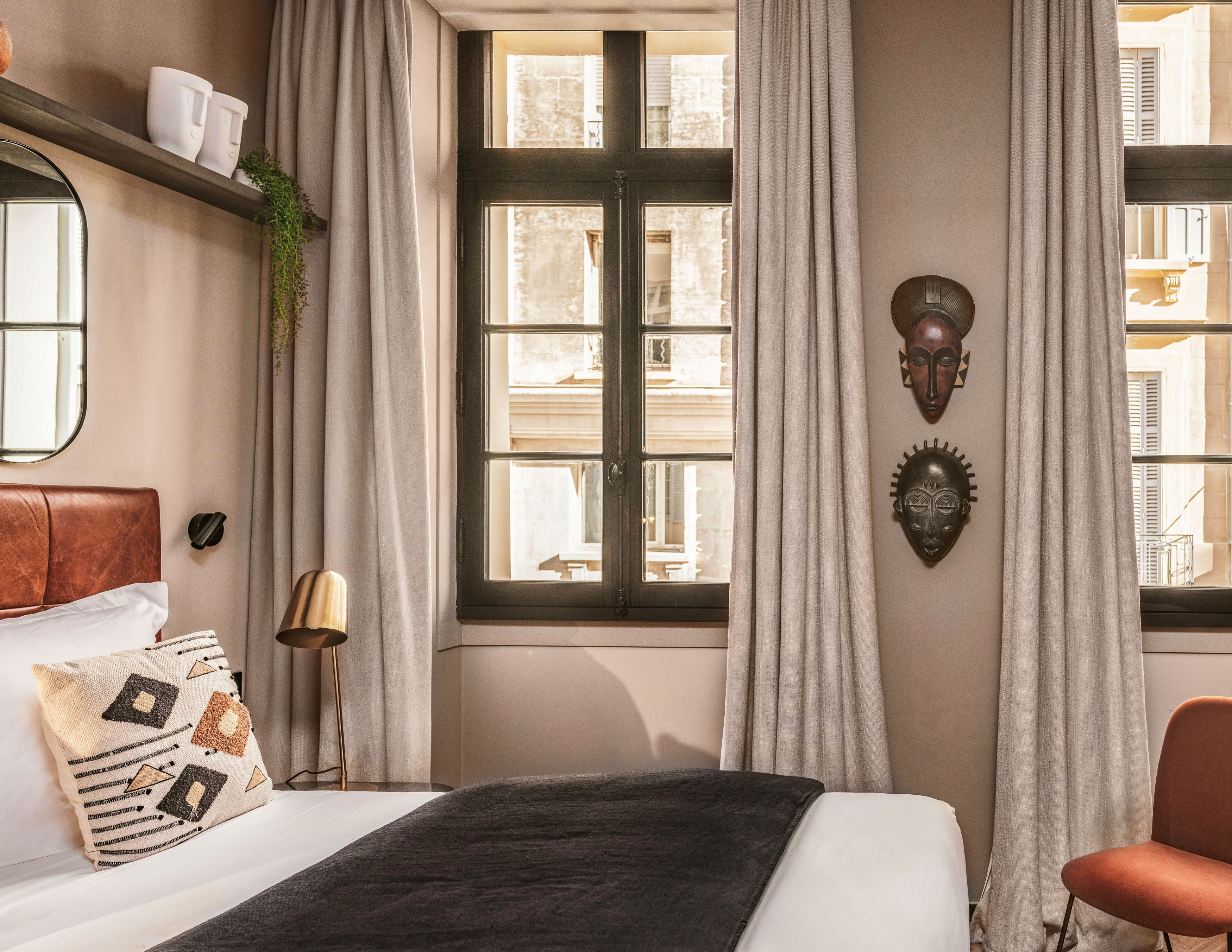A hotel room with a bed, a window with curtains, a wall-mounted light fixture, and a decorative mask.