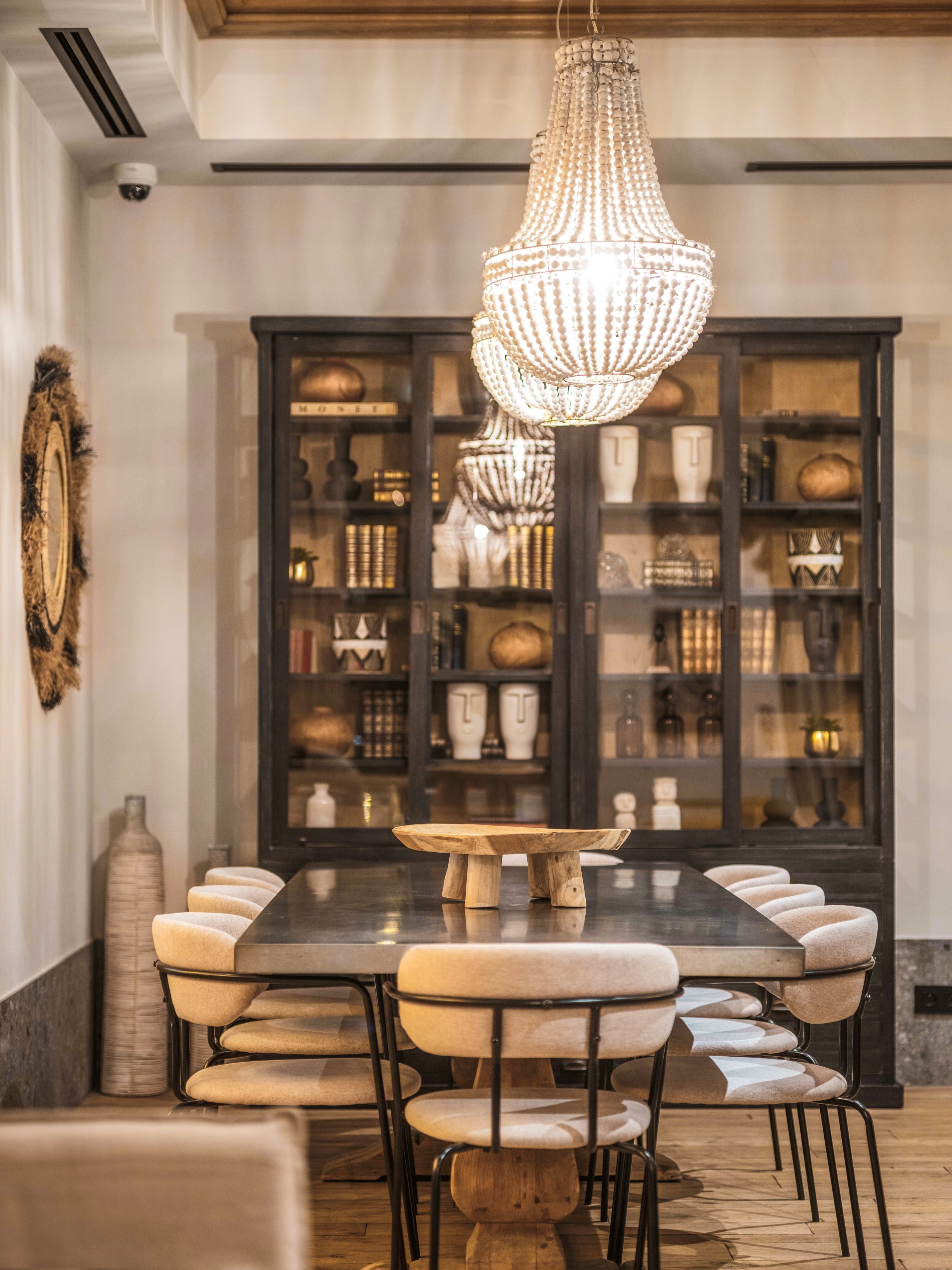A dining area with a large wooden table surrounded by upholstered chairs, a decorative chandelier, and a cabinet filled with various items.