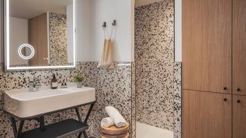 Une salle de bain moderne avec un lavabo blanc, un miroir avec éclairage intégré, un banc en bois et un espace douche.