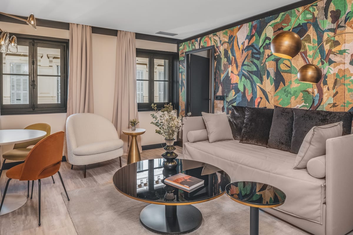 A modern living room with a sofa, coffee table, and chairs. There is a window with curtains, a colorful abstract wall, and a small table with a vase.