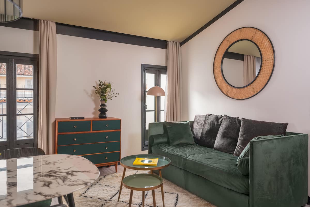 A living room with a green sofa, a round mirror, a small table with a book, a green cabinet, and a patterned rug.