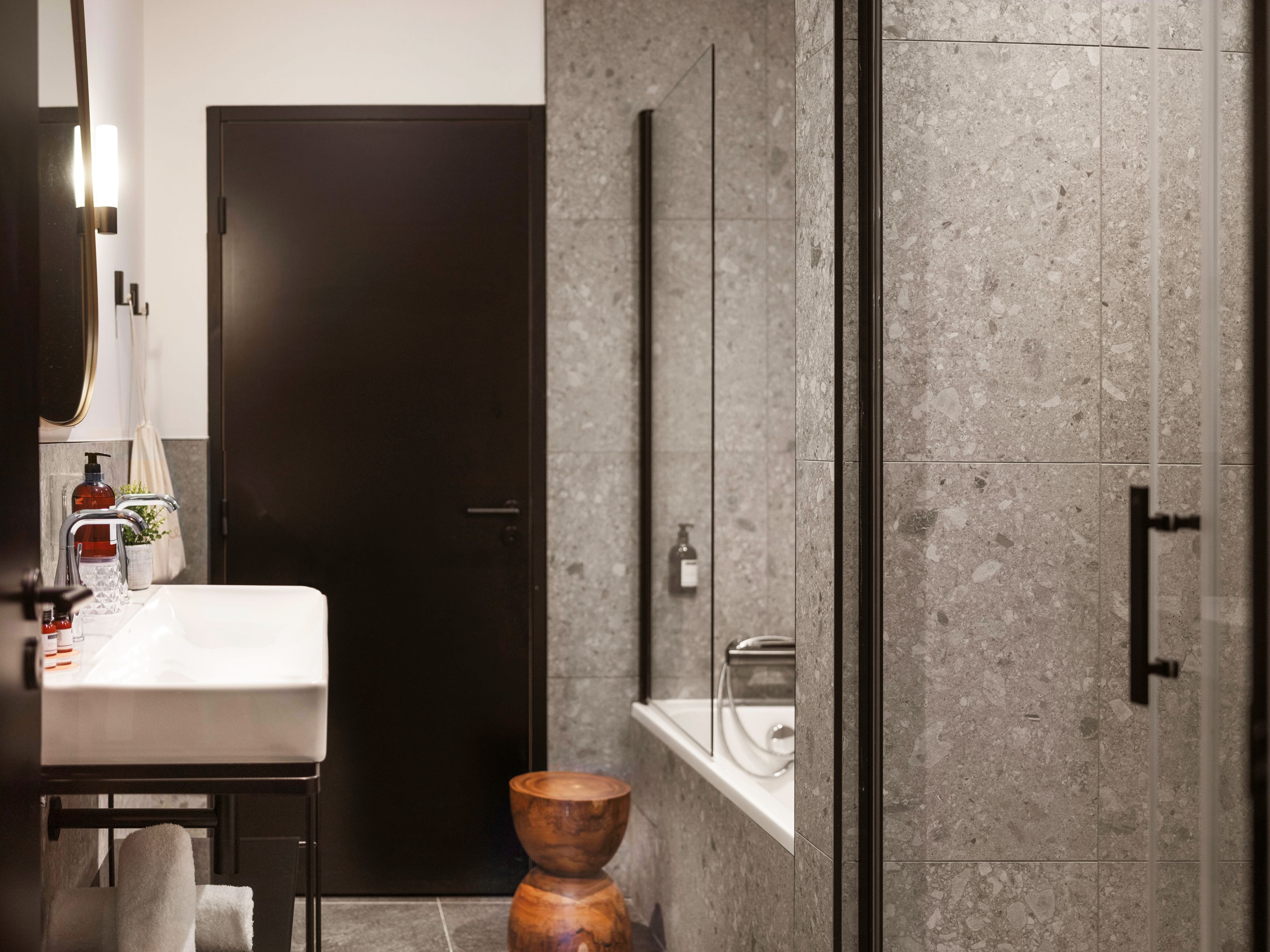 Een moderne badkamer met een donkere deur, een witte wastafel, een houten krukje en een douchecabine van glas.