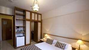 Habitación de hotel con cama, colcha a cuadros, dos lámparas de noche, mesitas de noche, escritorio con lámpara, refrigerador, armario con estantes y una puerta.