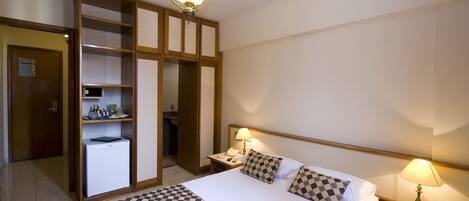 A hotel room with a bed, checkered bedspread, two bedside lamps, a nightstand, a desk with a lamp, a refrigerator, a wardrobe with shelves, and a door.