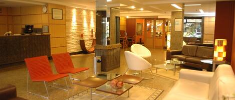 A hotel lobby with a reception desk, seating area with chairs and sofas, and a coffee machine.