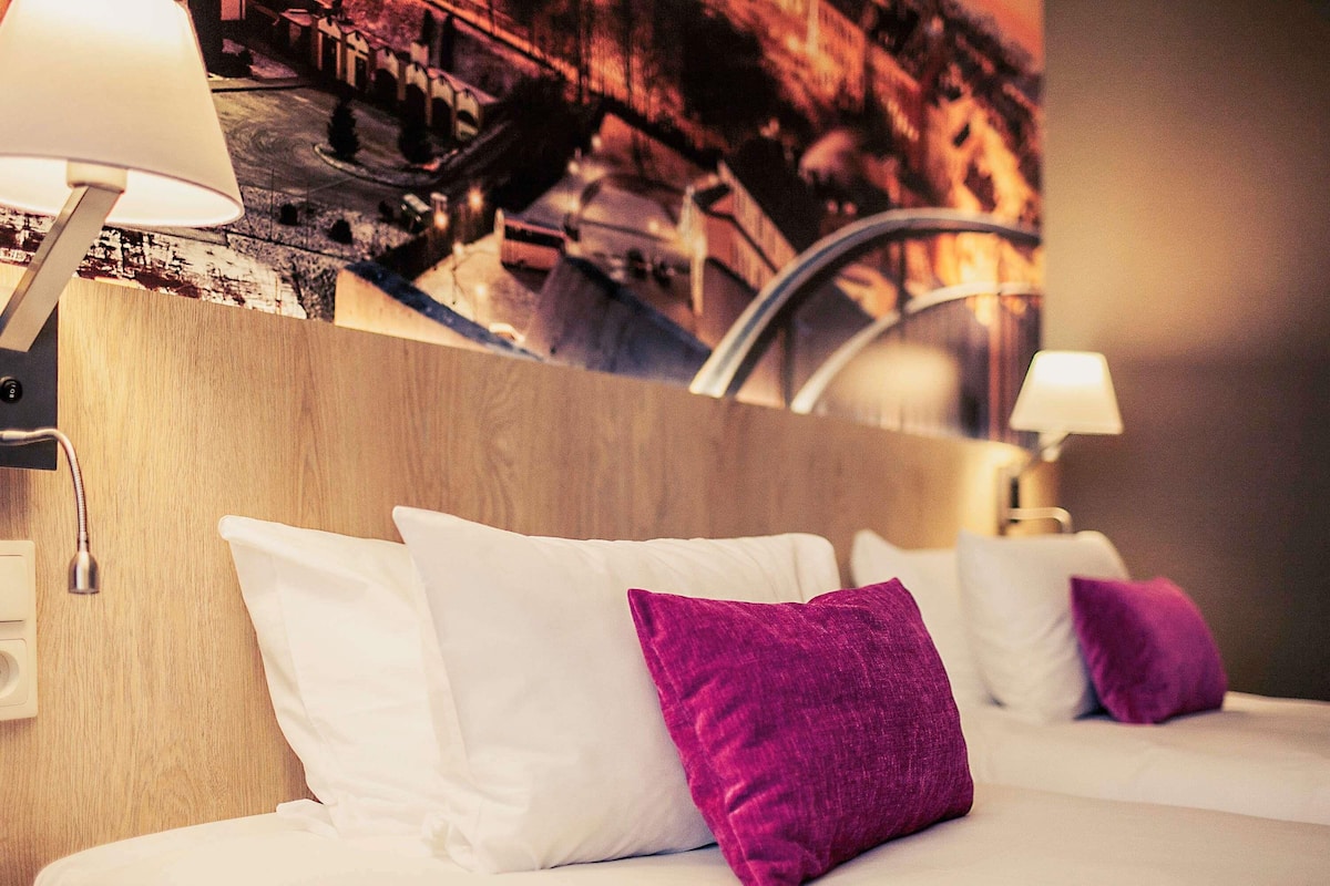 A hotel room with a bed, white and purple pillows, a wooden headboard, and a wall-mounted lamp.