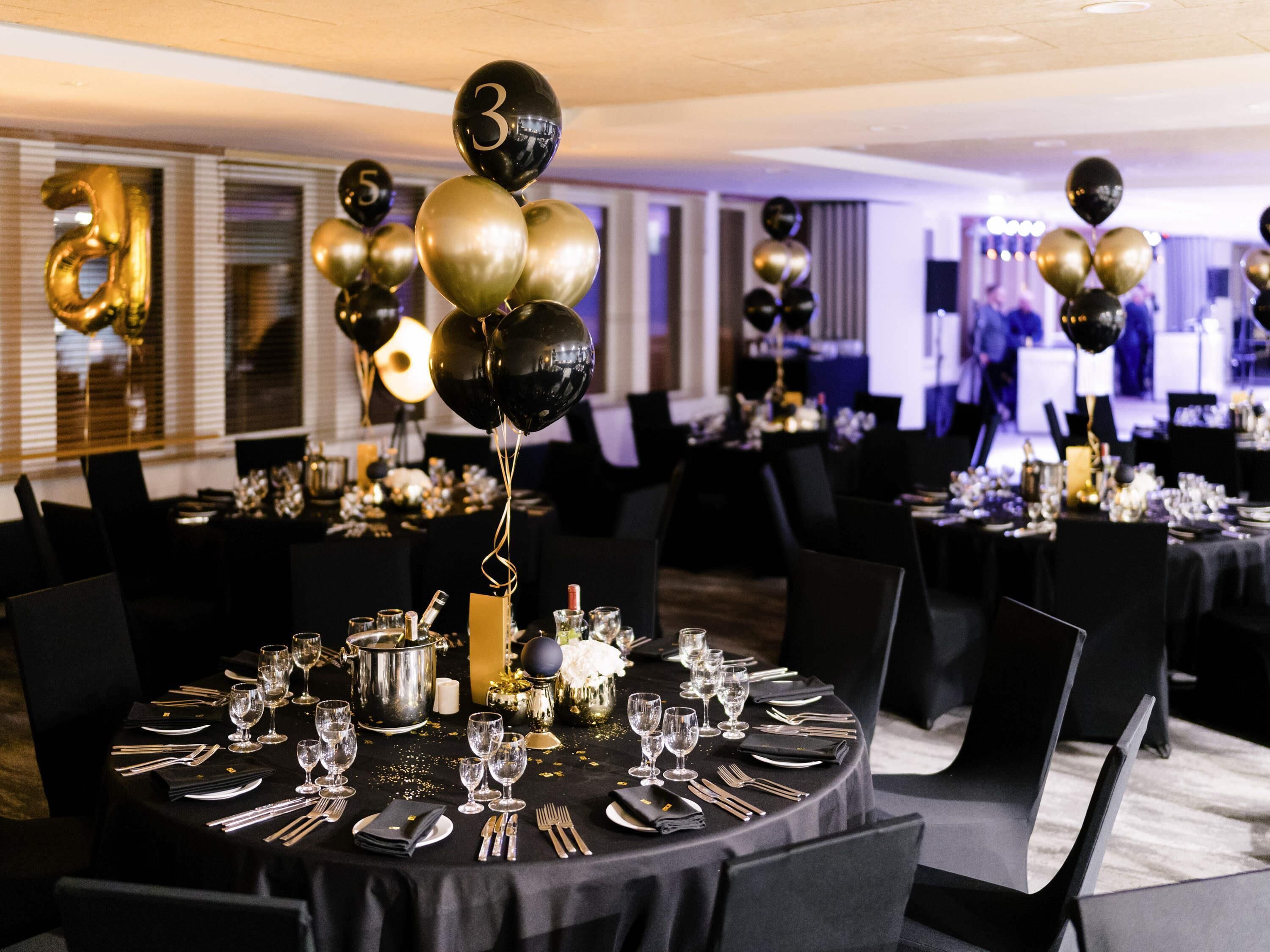 Un salón de banquetes con mesas redondas dispuestas para un evento formal, con decoraciones y globos dorados y negros.