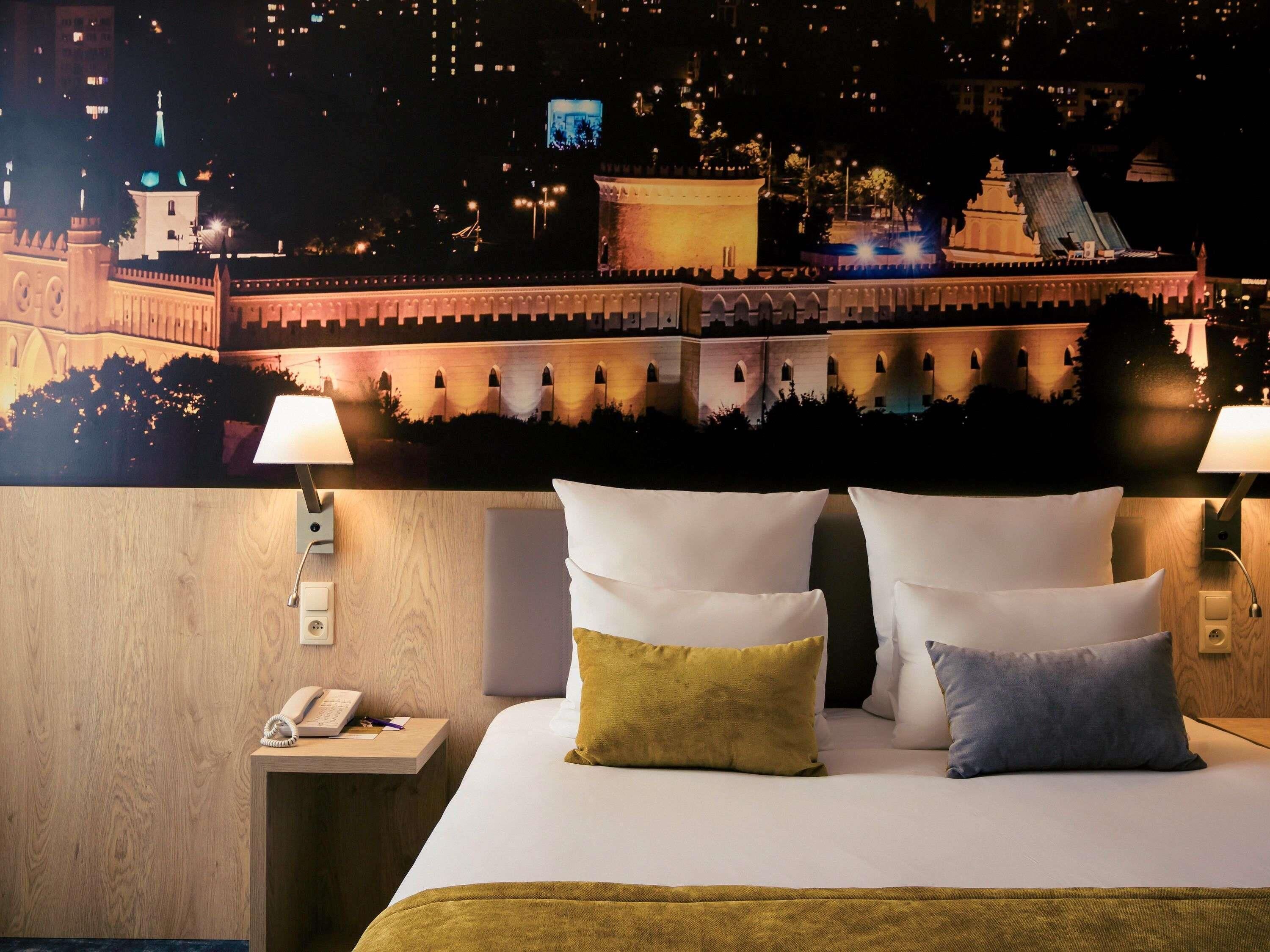 A hotel room with a bed, two bedside lamps, a nightstand with a telephone, and a wall mural of a cityscape.