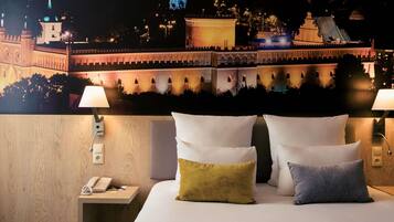 A hotel room with a bed, two bedside lamps, a nightstand with a telephone, and a wall mural of a cityscape.