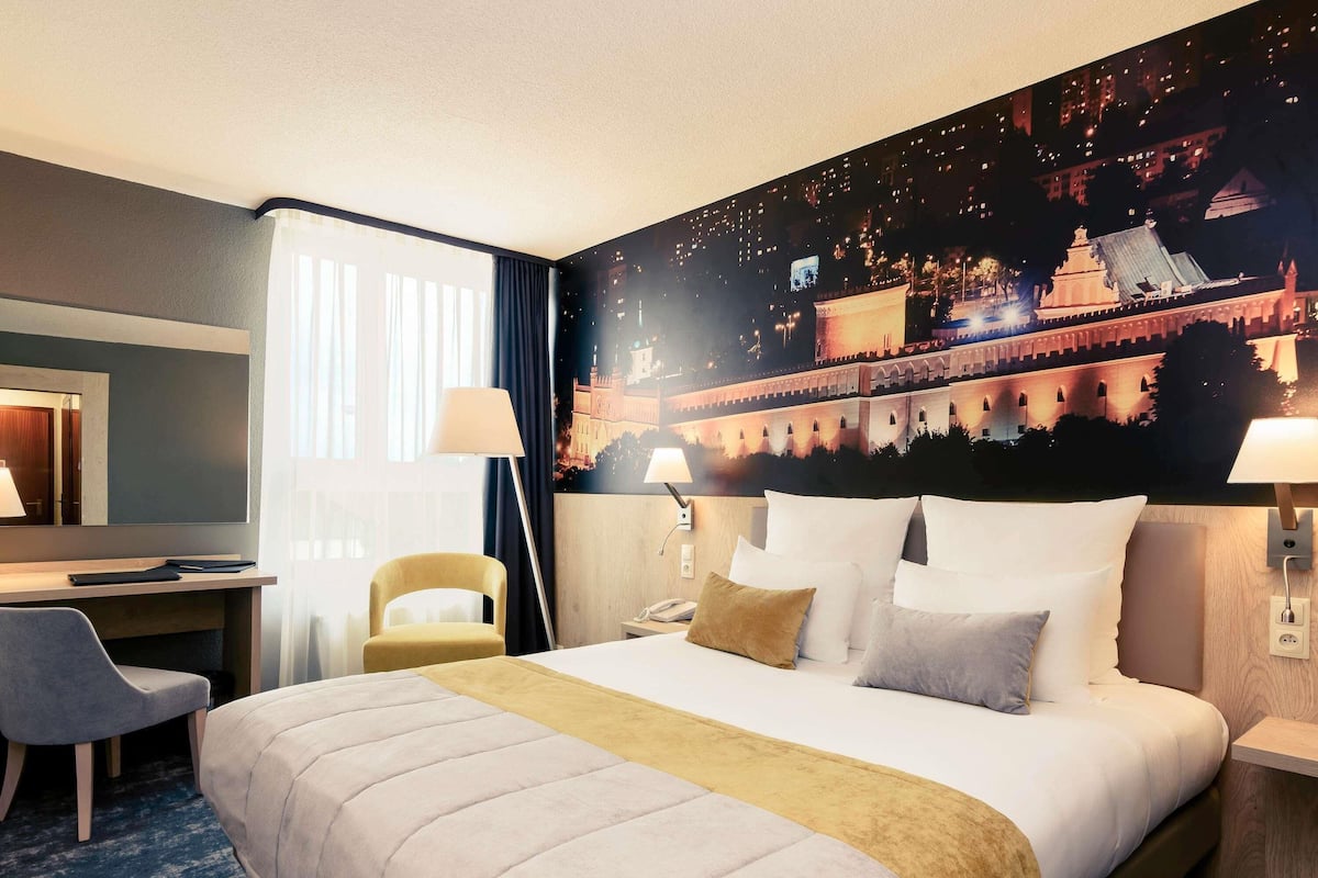 A hotel room with a bed, a desk, a chair, a lamp, and a wall mural.