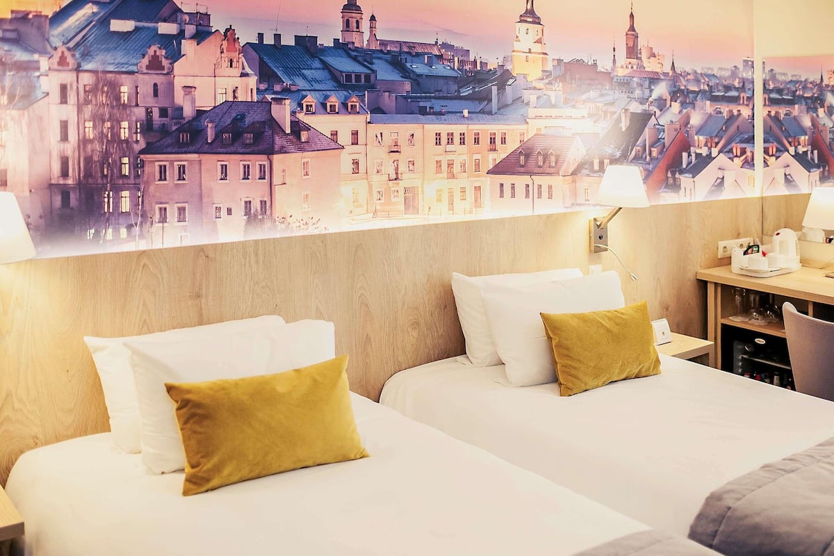 A hotel room with two beds, a wooden headboard, a nightstand with a lamp, and a cityscape view.