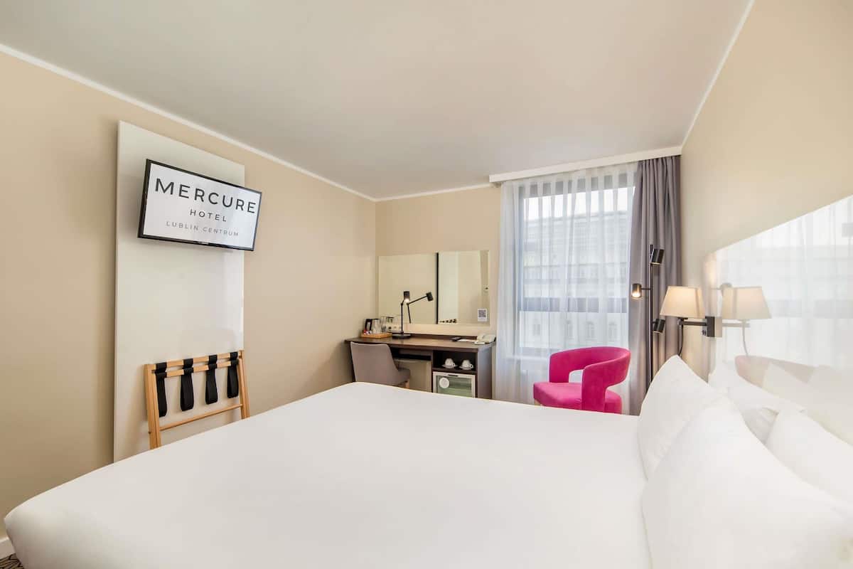 A hotel room with a bed, desk, chair, and a large window with curtains.