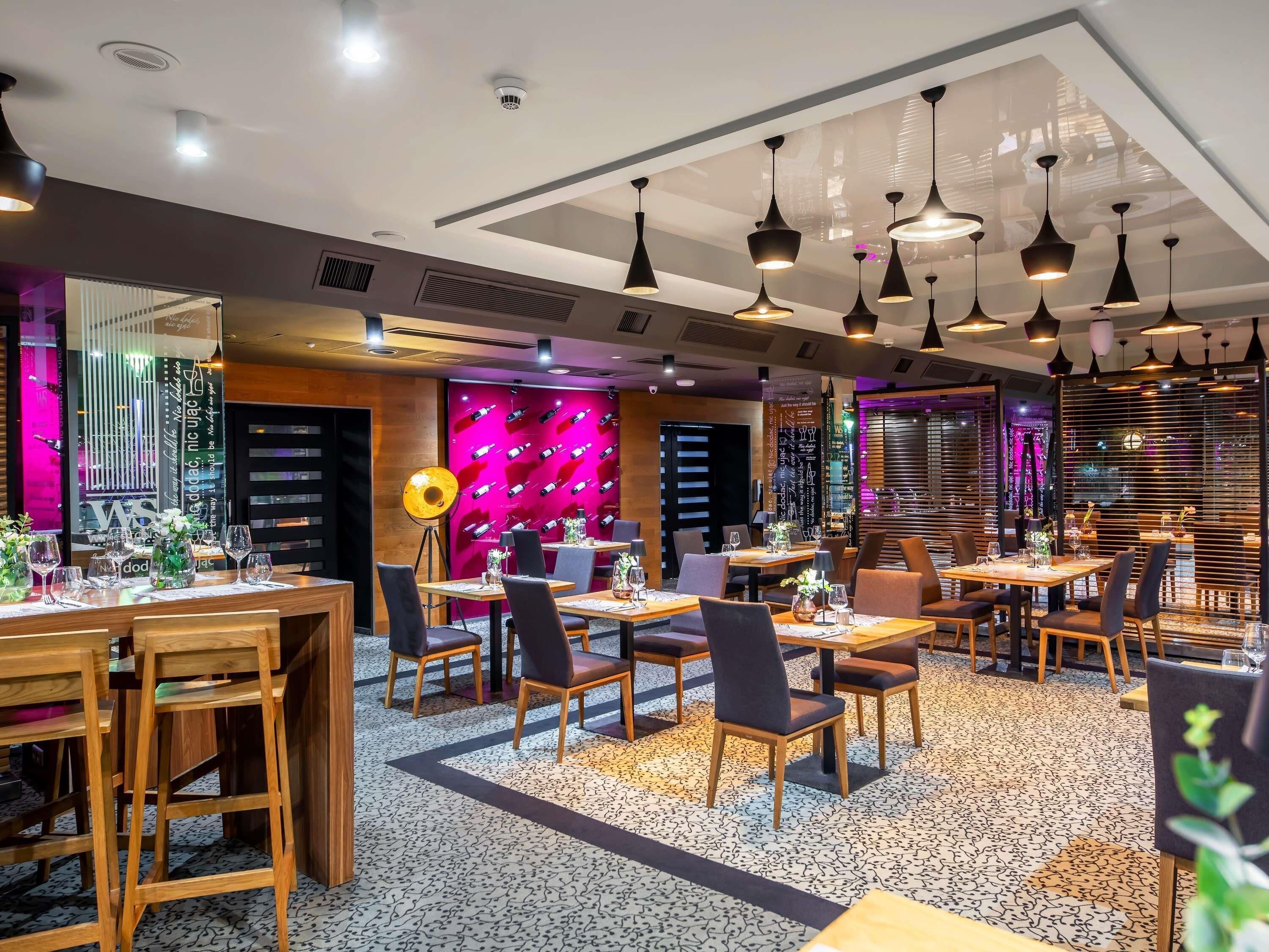 Ein modernes Restaurantambiente mit mehreren Esstischen, Stühlen, Pendelleuchten und einer lebendigen Wandgestaltung.